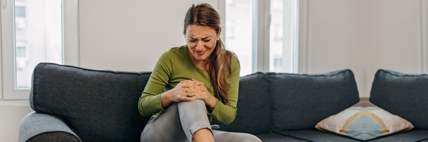Woman clutching knee with osteoarthritis and joint pain sitting on black couch