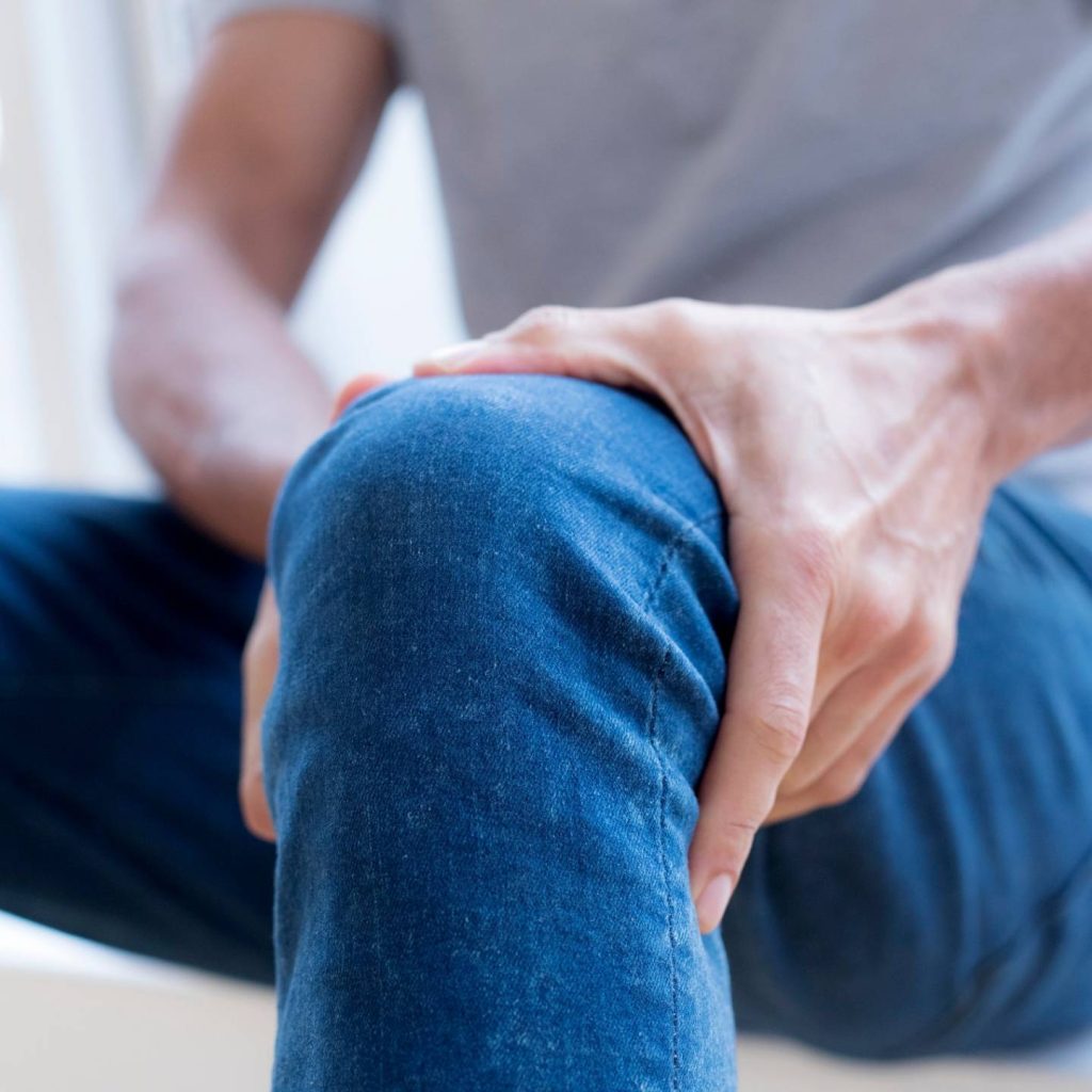 Man in grey shirt and blue jeans holding knee because of knee pain